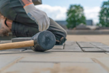 Close-up of a construction worker's gloved hands laying outdoor paving slabs on a prepared foundation on a summer sunny day, focus on a construction paving hammer