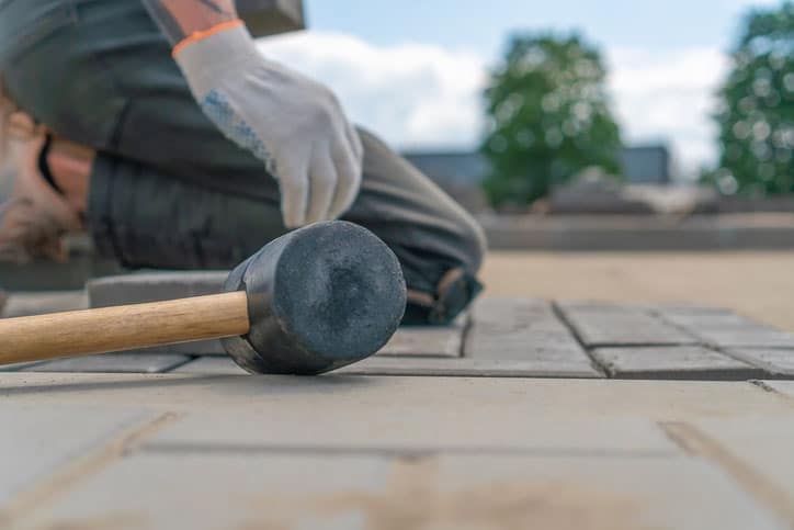 Close-up of a construction worker's gloved hands laying outdoor paving slabs on a prepared foundation on a summer sunny day, focus on a construction paving hammer