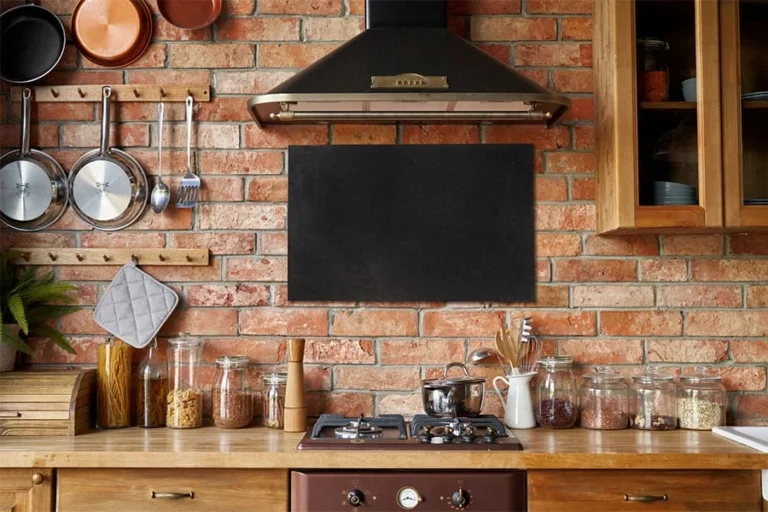 A cozy kitchen with a brick wall, hanging pots and utensils, a stove with a kettle, glass jars of ingredients on a wooden counter, and a black range hood above the stove—designed to blend charm and function in every detail.
