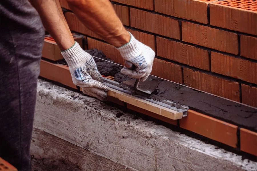 A person wearing white gloves uses a trowel to spread mortar between rows of red bricks, focusing on brick durability while constructing a sturdy brick wall. A person wearing white gloves uses a trowel to spread mortar between rows of red bricks, focusing on brick durability while constructing a sturdy brick wall.