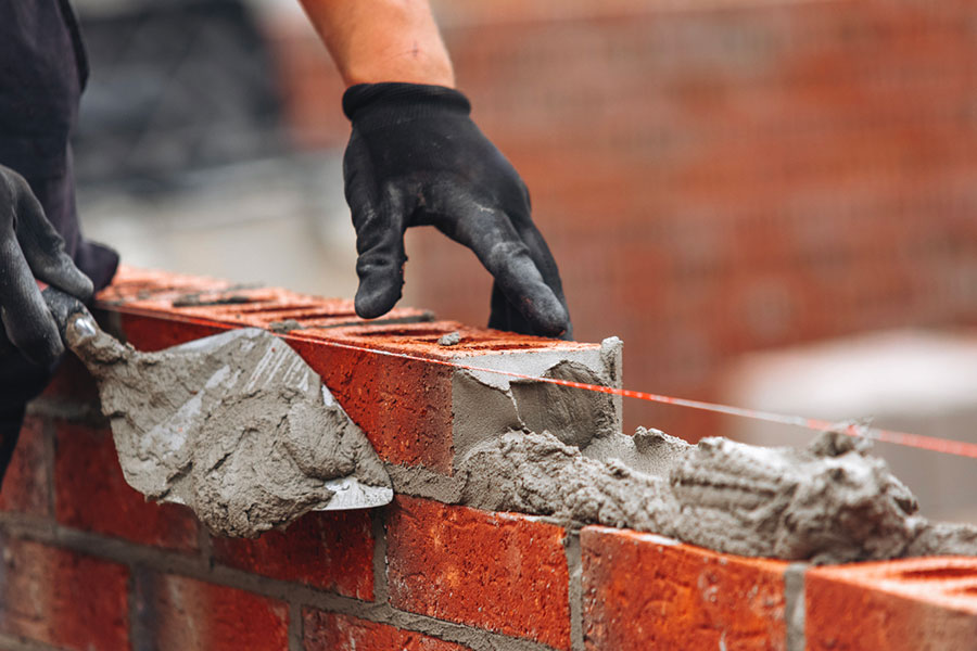 A person wearing black gloves spreads mortar on red bricks with a trowel, constructing a brick wall. Brick Contractors in Grosse Pointe, MI use a taut string line to ensure the bricks are laid straight.