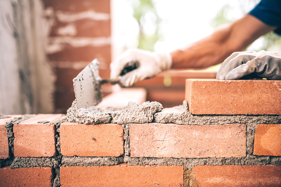A person wearing gloves uses a trowel to spread mortar while building a brick wall, with red bricks and wet cement clearly visible in the foreground—typical craftsmanship from skilled Brick Contractors in Michigan.
