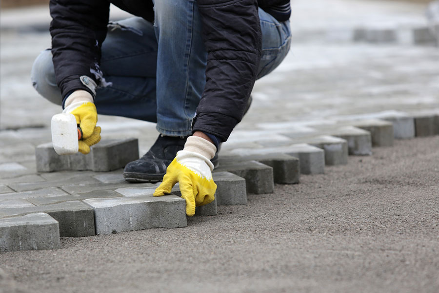 A person wearing yellow gloves and a dark jacket kneels on the ground, carefully laying gray paving stones in a row on a prepared sand or gravel surface—showcasing the skill of professional brick contractors.