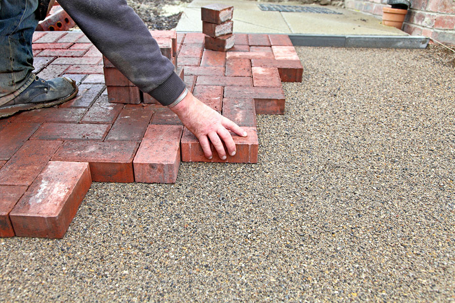 A person from Grosse Pointe, MI, lays red bricks on a flat gravel surface, creating a paved walkway or patio. Brick contractors' techniques are evident as their hand carefully places each brick, with stacked bricks visible nearby. A person lays red bricks on a flat gravel surface, creating a paved walkway or patio. Brick contractors' techniques are evident as their hand carefully places each brick, with stacked bricks visible nearby.
