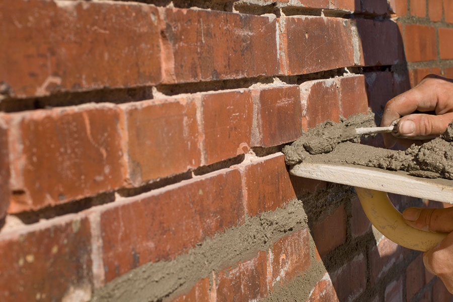 A close-up of a person applying fresh mortar between red bricks on a wall with a trowel, showcasing skilled masonry repair.