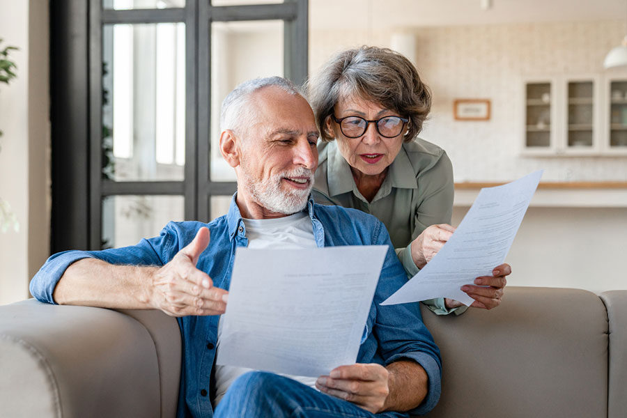 An older man and woman sit together on a couch, discussing documents about brick installation cost. The man smiles while holding a paper, as the woman, standing behind him, points to details on another sheet.