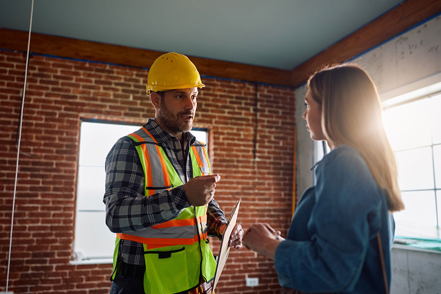 A construction worker in a hard hat and safety vest talks to a woman inside a partially finished brick building, clipboard in hand, as sunlight streams through the window—highlighting why Michigan homeowners hire masonry contractors.