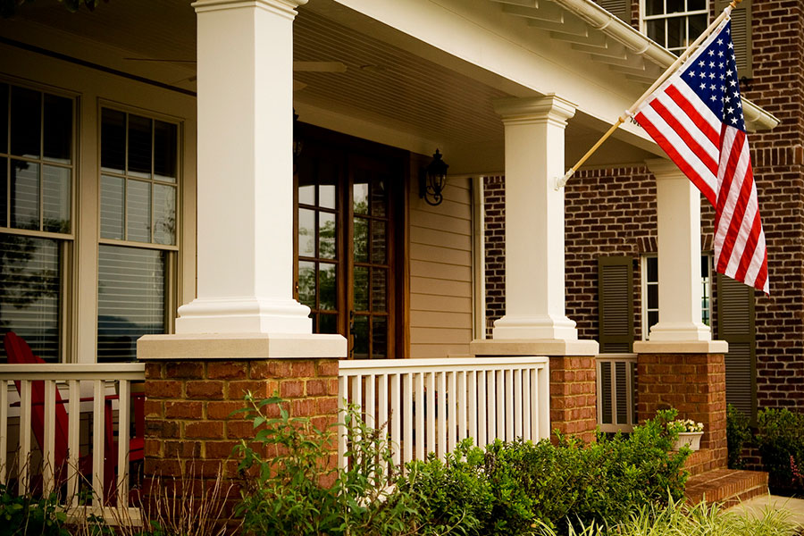 American flag hanging from the front porch of a brick house with white pillars, large windows, a wooden door, and greenery in the foreground—an inviting scene that highlights the charm of a covered brick porch.