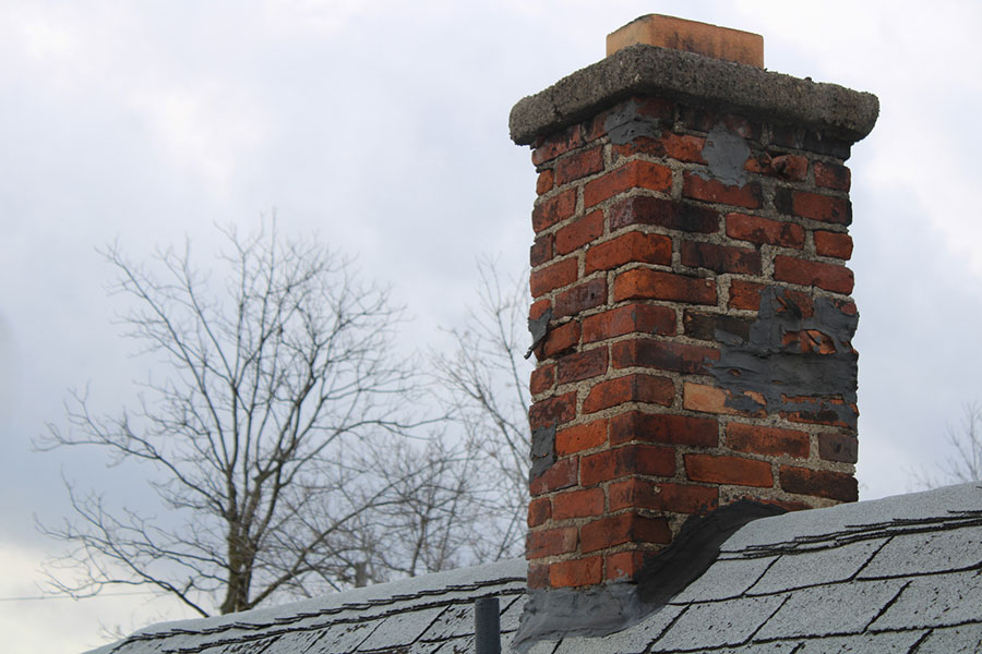 A weathered red brick chimney with some loose chimney bricks rises from a shingled rooftop, with leafless trees and a cloudy sky in the background.