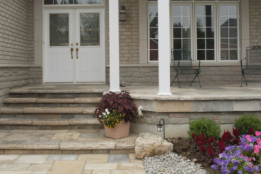 Front porch with stone steps, white double doors, a large potted plant, black metal chairs, and a small garden with flowers, shrubs, rocks, and mulch—this inviting front patio adds charming curb appeal to any Michigan home.