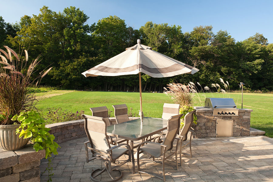 An outdoor brick patio featuring a glass-topped dining table, six chairs, and a striped umbrella next to a built-in stone grill.