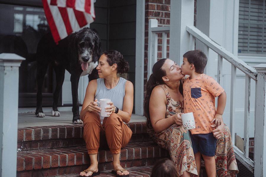 Two women and a young boy sit on a brick porch, with one woman kissing the boy’s cheek while a large dog stands nearby, all gathered safely on the wide steps despite the uneven steps leading up.